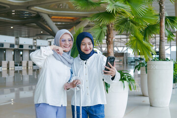 Two Asian women in hijab smiling while making a video call with relatives at the airport, sharing joyful moments before departure