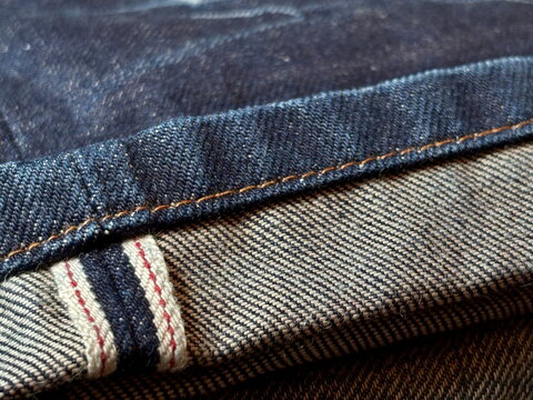 Close-up macro of selvedge denim cuff with orange stitching, showing detailed twill texture for fashion design, craft and textile backgrounds.