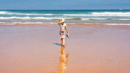 Tourist woman in dress and hat strolling along a lonely golden sand beach in northern Spain, Costa Quebrada, Cantabria.