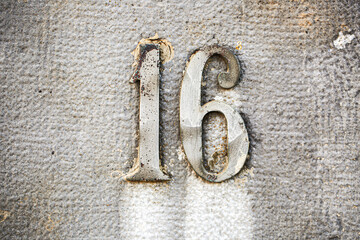 Number 16 in vintage metal numerals mounted on a textured concrete wall. Weathered and rustic...
