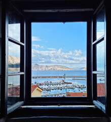 A picturesque view through a dark-framed window reveals the vibrant Baska harbor filled with boats, the glistening Adriatic Sea, and the distant outline of Prvic Island under a bright blue sky