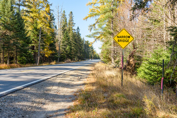 Yellow traffic sign warning against a narrow bridge along a forest road in the mountains on a clear autumn day