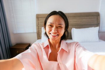 Smiling asian young female in pink shirt taking a selfie at home