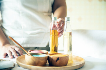 Hand selecting a glass bottle of essential oil beside colorful spa salts in wooden bowls on tray; wellness preparation for therapeutic massage and aromatherapy treatment