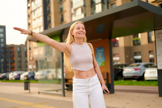 smiling woman hailing taxi at bus stop in urban area, smartphone in hand. casual summer outfit, sunny day. modern cityscape background, transportation lifestyle concept.