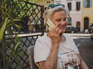 Senior woman speaking on smartphone, wearing sunglasses atop white hair, seated outside residential...