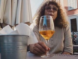 Mature woman wearing sunglasses relaxing at outdoor cafe, sipping white wine, basking in sunlight with contemplative expression