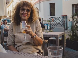 Smiling mature woman with curly hair, glasses, sipping spritz cocktail while relaxing at sunlit...