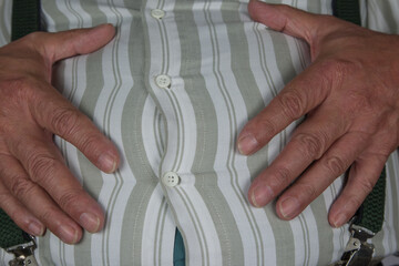 A man with shirt buttons stretched to the limit, about to burst. His hands rest on his full belly, a clear sign of overeating and excess weight.