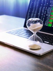 Hourglass standing on laptop, stock market quotes on screen in background.