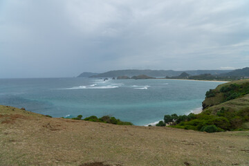 Obraz premium Scenic ocean view in Lombok, Indonesia. Waves crash on rocky islets with turquoise water and lush coastline under a cloudy sky