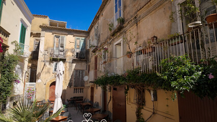 Tables and chairs arranged beneath the balconies of weathered buildings, creating a quaint dining...