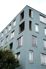 Contemporary residential building with blue-gray facade and multiple windows under cloudy sky. Green tree and parking sign in foreground. Urban apartment architecture with clean lines.
