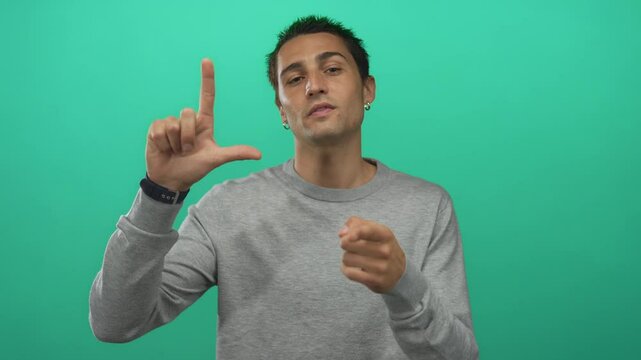 Hispanic man with defiant stare makes loser sign and points finger to camera in studio; mockery teasing.