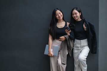 Two young businesswomen using smartphone and holding laptop near gray wall