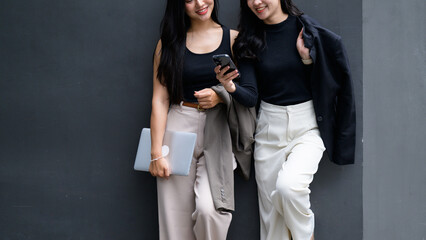 Two businesswomen are using a smartphone and holding a laptop near a gray wall