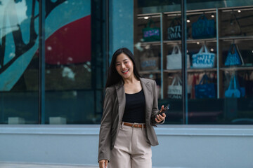 Smiling businesswoman walking and using smartphone in front of modern building