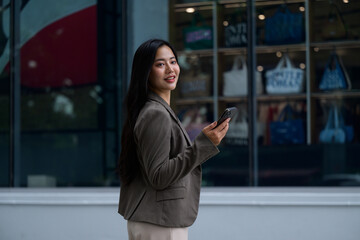 Young Asian businesswoman using smartphone while walking near shop window