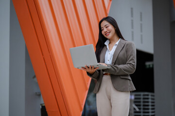 Smiling businesswoman using laptop standing near modern orange building