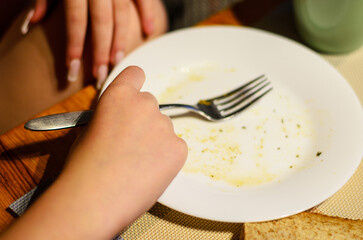 Child hand holding fork over empty plate on wooden dining table indoors at home