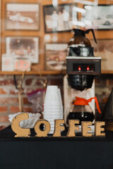 Rustic coffee station with vintage wooden COFFEE sign, stacked paper cups, commercial coffee brewer, warm indoor brick wall with framed photographs in cozy café or event setting
