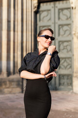 Portrait young adult elegant woman black dress and sunglasses smiles confidently in golden sunlight warm sunset backlit light against historic cathedral building. Female person walking city street