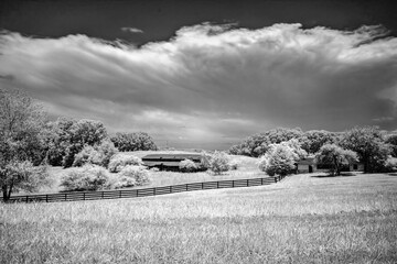 This striking black-and-white infrared photograph captures a rustic barn nestled in a serene countryside field, surrounded by lush, glowing trees under a dramatic sky , evoking timeless tranquility .