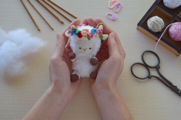 Crocheted sheep toy with floral crown held by female hands after completion DIY amigurumi soft...