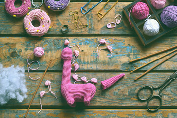 Amigurumi dinosaur toy made of pink yarn with legs and tail visible surrounded by wool balls and...