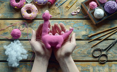 Hands holding pink amigurumi dinosaur body with yarn and stuffing preparing for final assembly of...