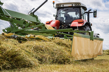 Large red tractor moving green farmers pasture, motion close up. Big red tractor collect the grass for silage. Agricultural machine working during harvest.