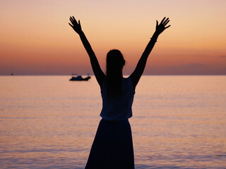 Silhouette of a woman standing on the beach with arms raised, enjoying the peaceful sunset over the sea. Calm ocean waves, pastel sky colors, summer mood and relaxing vacation atmosphere