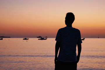 Silhouette of a man watching the sunset at the beach and enjoying the peaceful sea view. Tranquil mood, calm water, colorful sky and relaxing summer scene
