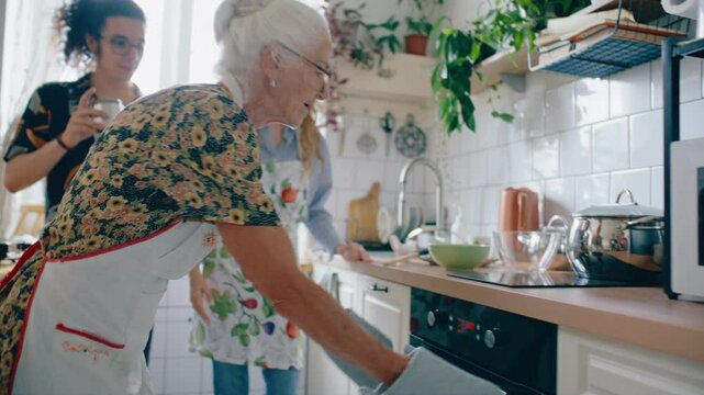 Elderly grandmother in apron and mitts putting baking tray of homemade pizza into hot oven while cooking with family in kitchen