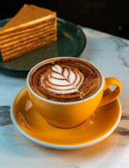 Hot Mocha with Leaf Latte Art in Yellow Cup on Marble Table. Blurred slice of layered dessert in background. Ideal for menus, cafes, cozy lifestyle visuals, and branding.
