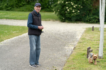 Elderly man in a baseball cap walking a small dog on a leash in a summer park. Concept: active...