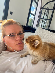 Woman with glasses relaxing on a couch, interacting with a small fluffy dog, showcasing a cozy...