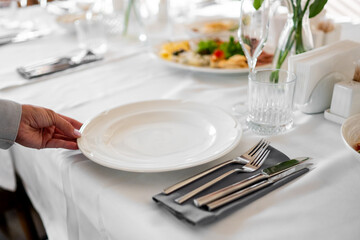 Hand placing a white plate on a neatly set dining table with cutlery, glass, and food in the...