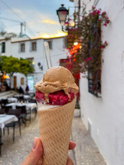 Delicious ice cream cone featuring rich flavors of chocolate and raspberry, held in hand against a charming street with blooming flowers and cozy outdoor seating