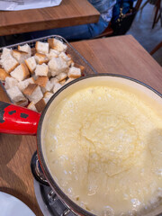 Creamy cheese fondue served in a metal pot with a red handle, accompanied by toasted bread cubes on a wooden table, inviting a delightful dining experience
