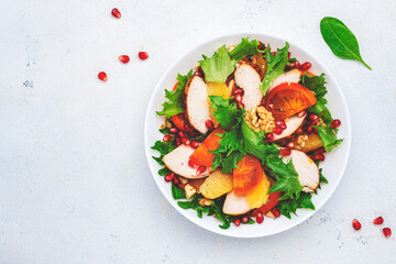 Autumn persimmon salad with grilled chicken fillet, pomegranate seeds, walnuts, oranges and lettuce, white table background, top view