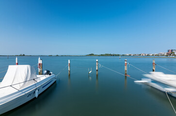 Wooden pier over calm lagoon waters in Grado, Italy, with boats moored under a bright summer sky and a peaceful view toward the horizon.