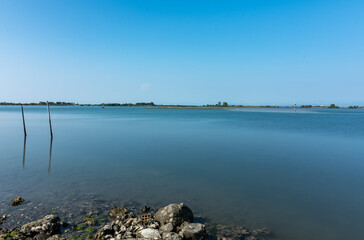 Peaceful lagoon view in Grado, Italy, with calm waters, a stony shoreline, and distant boats under a bright summer sky.
