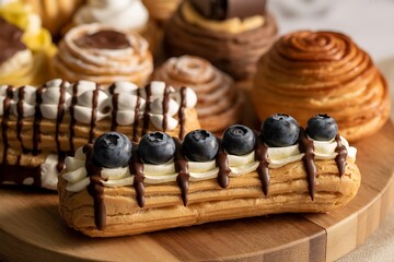 A delectable eclair pastry is topped with smooth cream, chocolate drizzle, and fresh blueberries on a wooden board
