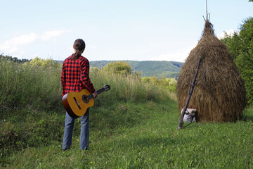 A young guy in a red shirt and jeans with a guitar travels alone through green mountains and nature.
