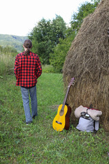 A young guy in a red shirt and jeans with a guitar travels alone through green mountains and nature.