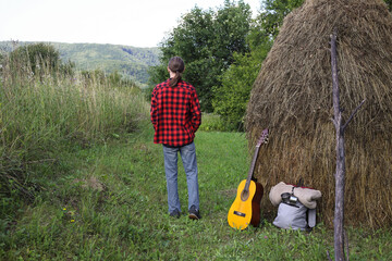 A young guy in a red shirt and jeans with a guitar travels alone through green mountains and nature.