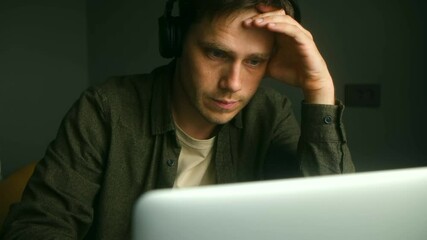 Young professional experiencing work related stress while listening to music, sitting at laptop with hand on head, displaying mental exhaustion and professional pressure during remote work session