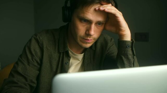 Young professional experiencing work related stress while listening to music, sitting at laptop with hand on head, displaying mental exhaustion and professional pressure during remote work session