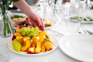 A hand reaches for a plate of assorted fresh fruits—grapes, oranges, strawberries, apples, and pears—on a bright white table set for a colorful and healthy meal.